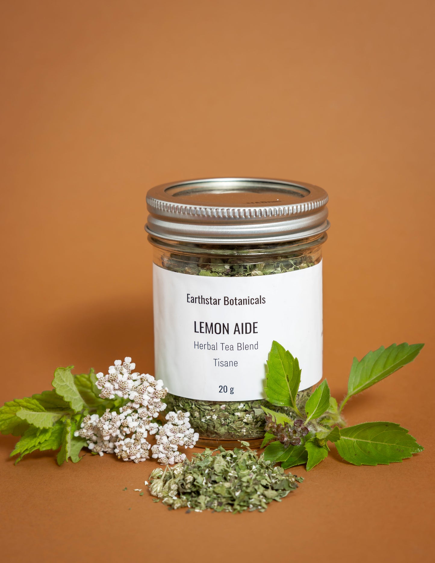 Jar of Earthstar Botanicals Lemon Aide Herbal Tea Blend on a brown background with herbs and flowers.