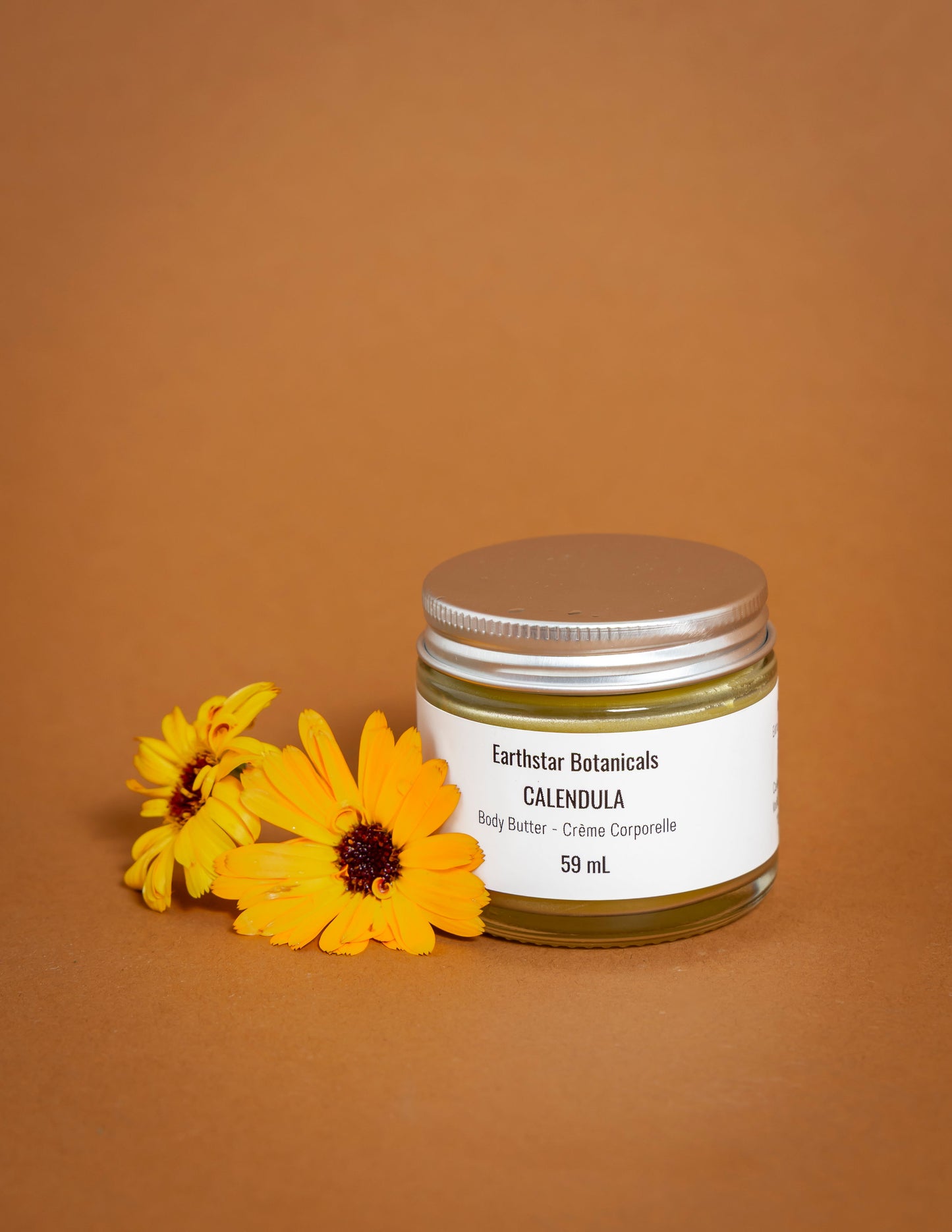 Jar of Earthstar Botanicals Calendula body butter with yellow Calendula flowers on an orange background