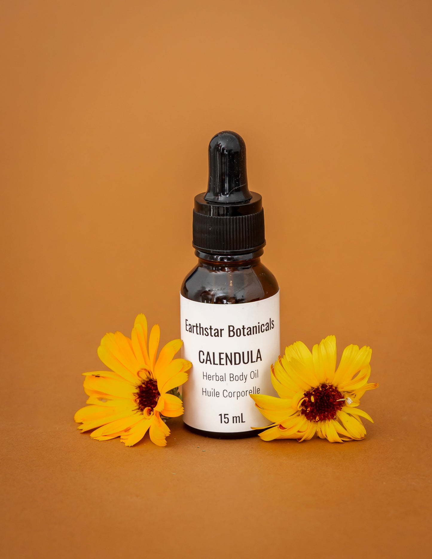 Bottle of Earthstar Botanicals Calendula Herbal Body Oil with two golden Calendula flowers on an orange background