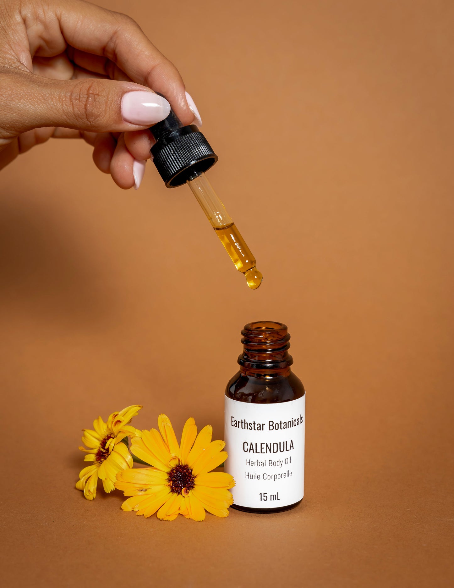 Hand holding a dropper with a bottle of Earthstar Botanicals Calendula Herbal Body Oil on an orange background.