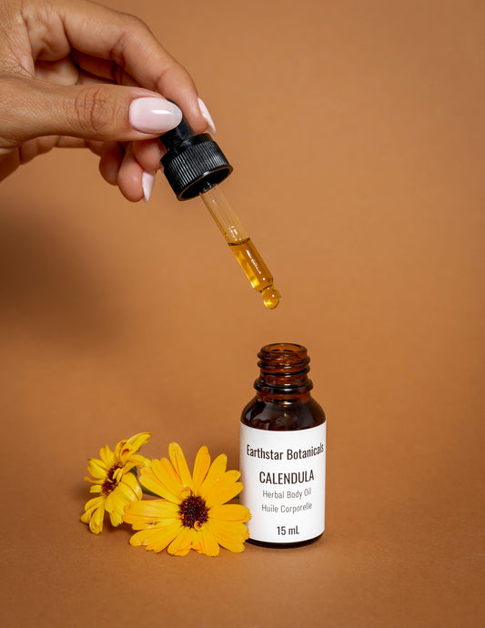 Hand holding a dropper with a bottle of Earthstar Botanicals Calendula Herbal Body Oil on an orange background.