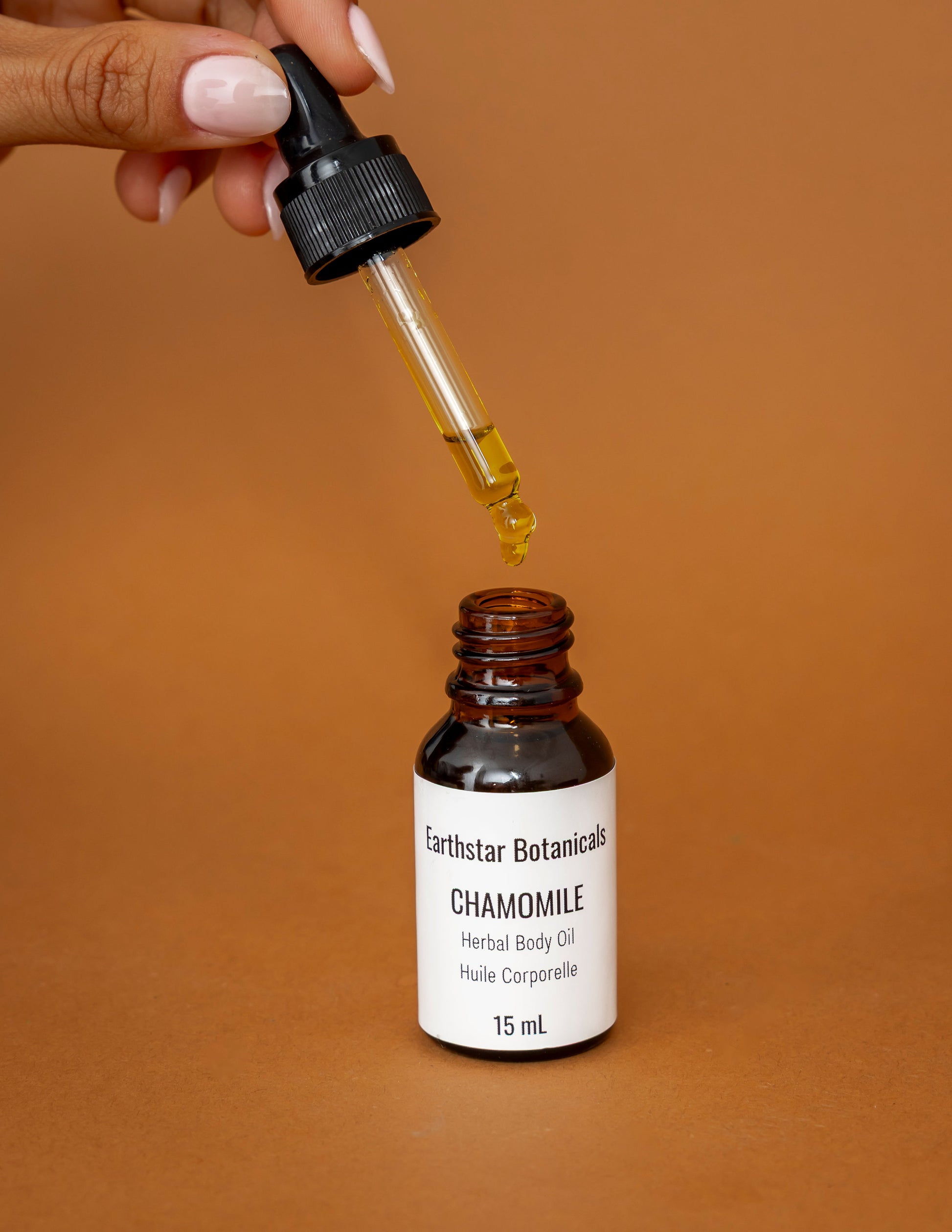 Chamomile Herbal Body oil being dripped from a dropper into a brown bottle with a white label on an orange background.