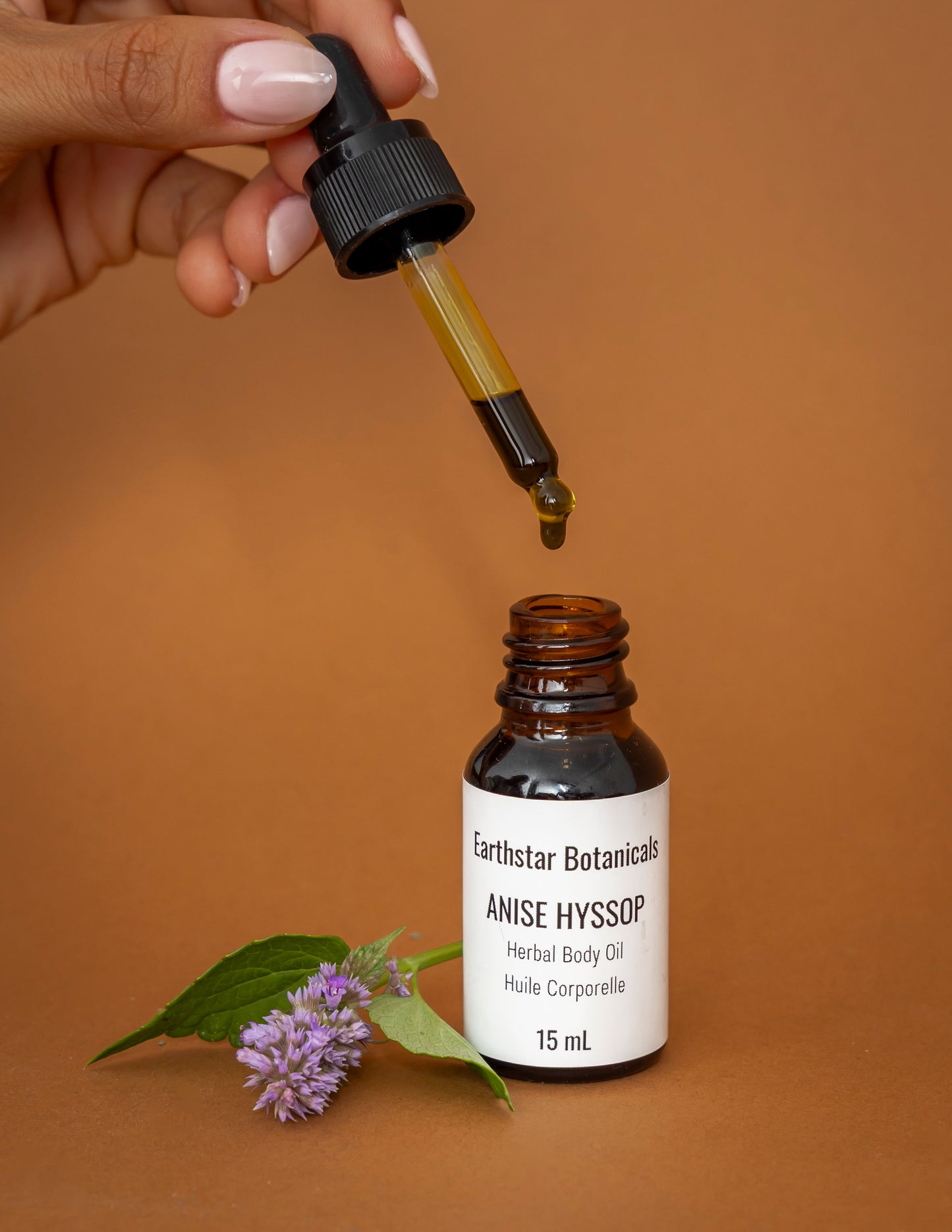 Hand holding a dropper over a bottle of Earthstar Botanicals Anise Hyssop Herbal Body Oil on an orange background.
