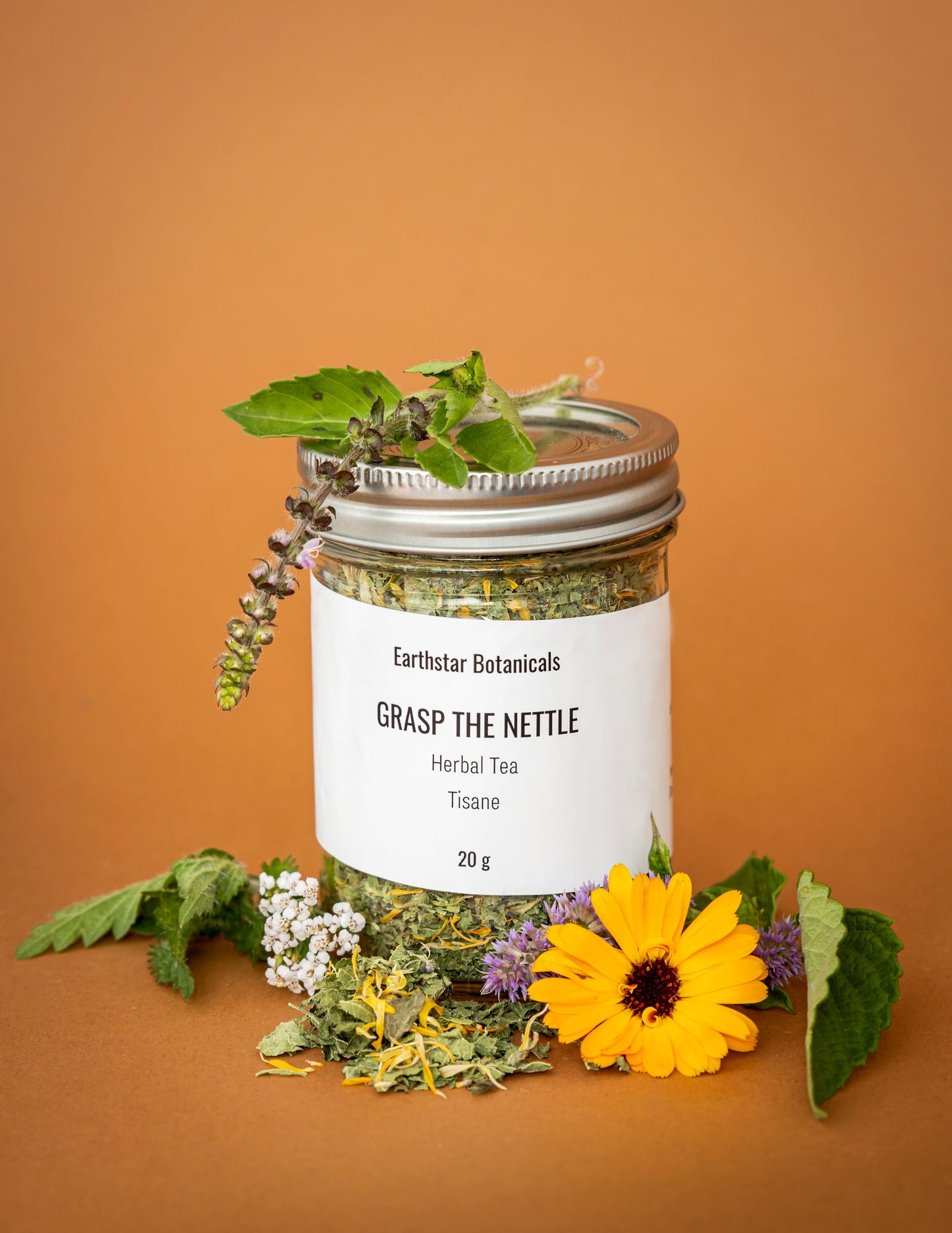 Jar of Earthstar Botanicals 'Grasp the Nettle' herbal tea surrounded by Calendula, Anise Hyssop, and Yarrown flowers and Besobela Tulsi leaves on an orange background