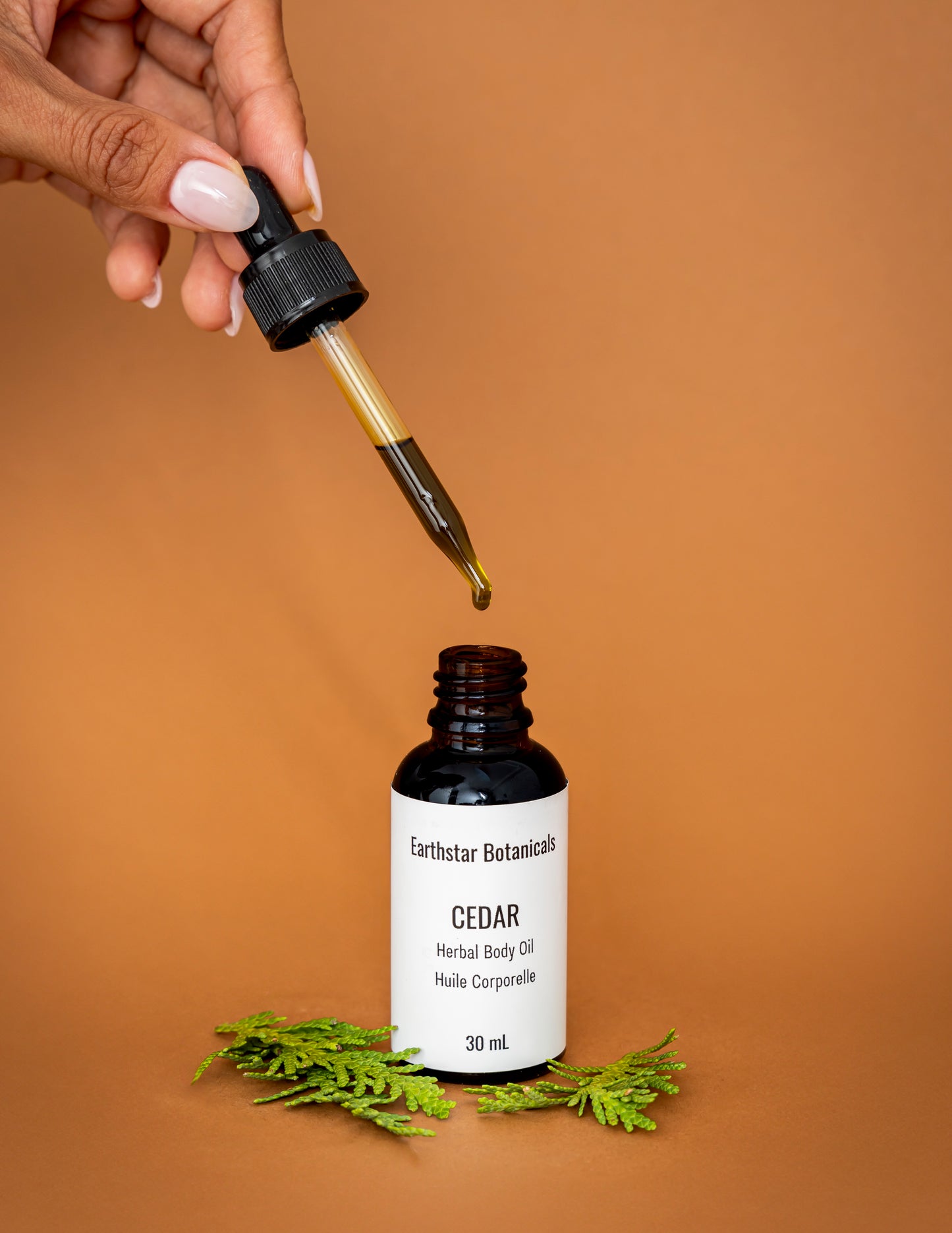 Hand holding a dropper over a bottle of Earthstar Botanicals Cedar Herbal Body Oil on an orange background.